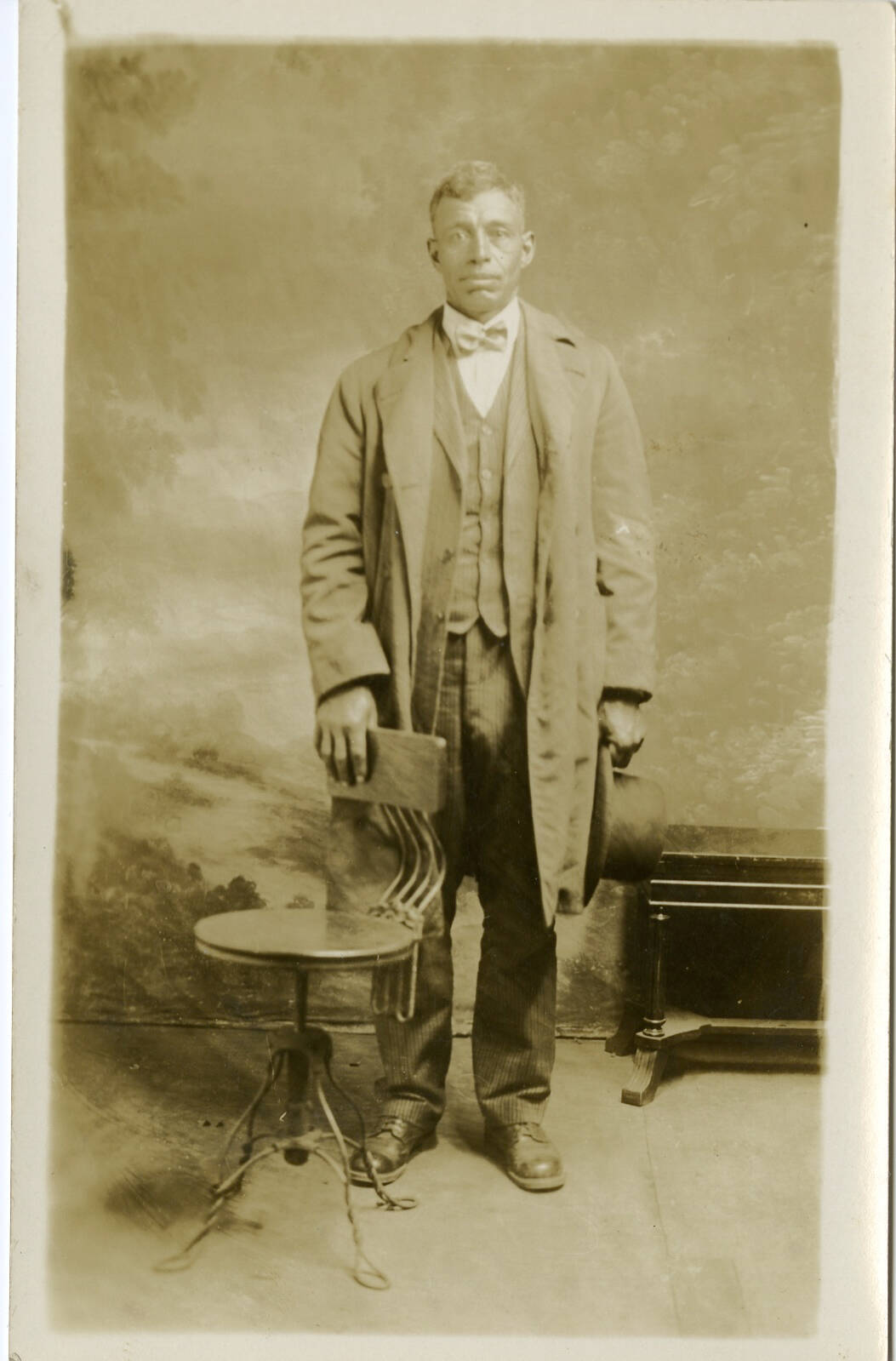 Photographic portrait of a man wearing a long jacket and suit, one hand on the back of a chair in front of him